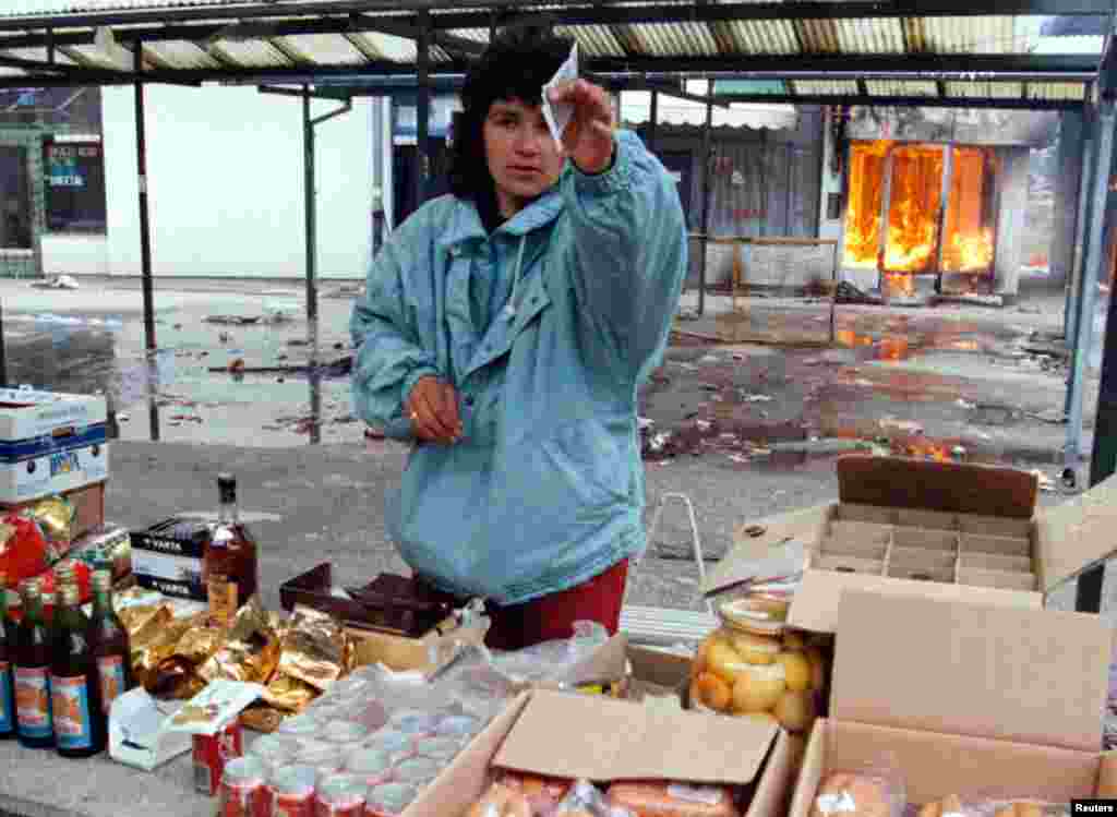 Serb aýal bazarda zat satýar, onuň arkasynda Ilidziada, Saraýewonyň serbleriň elindäki eteginde ýanýan dükan görünýär. 9-njy mart, 1996. &nbsp;
